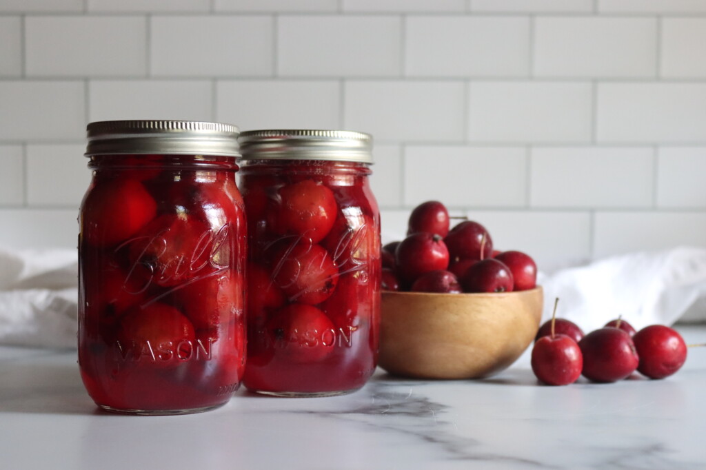 Two pint jars of home canned crabapples on a white counter with a bowl of crabapples next to them.