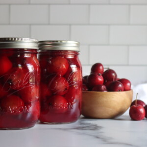 Two pint jars of home canned crabapples on a white counter with a bowl of crabapples next to them.