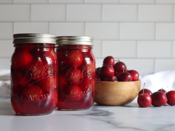 Two pint jars of home canned crabapples on a white counter with a bowl of crabapples next to them.