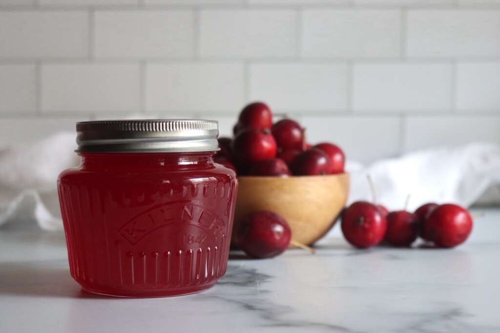 A jar of homemade crabapple jelly sitting in front of a bowl of red crabapples.