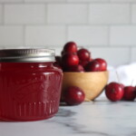 A jar of homemade crabapple jelly sitting in front of a bowl of red crabapples.