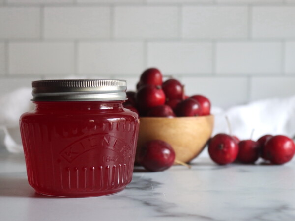 A jar of homemade crabapple jelly sitting in front of a bowl of red crabapples.