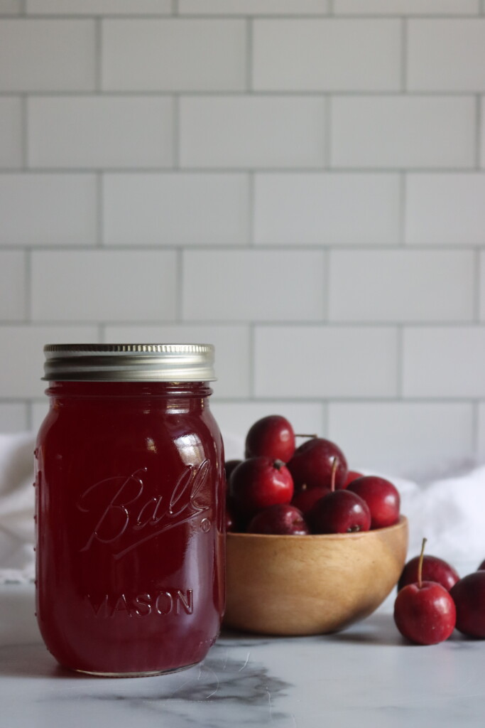 A jar of crabapple juice on a table next to red crabapples.