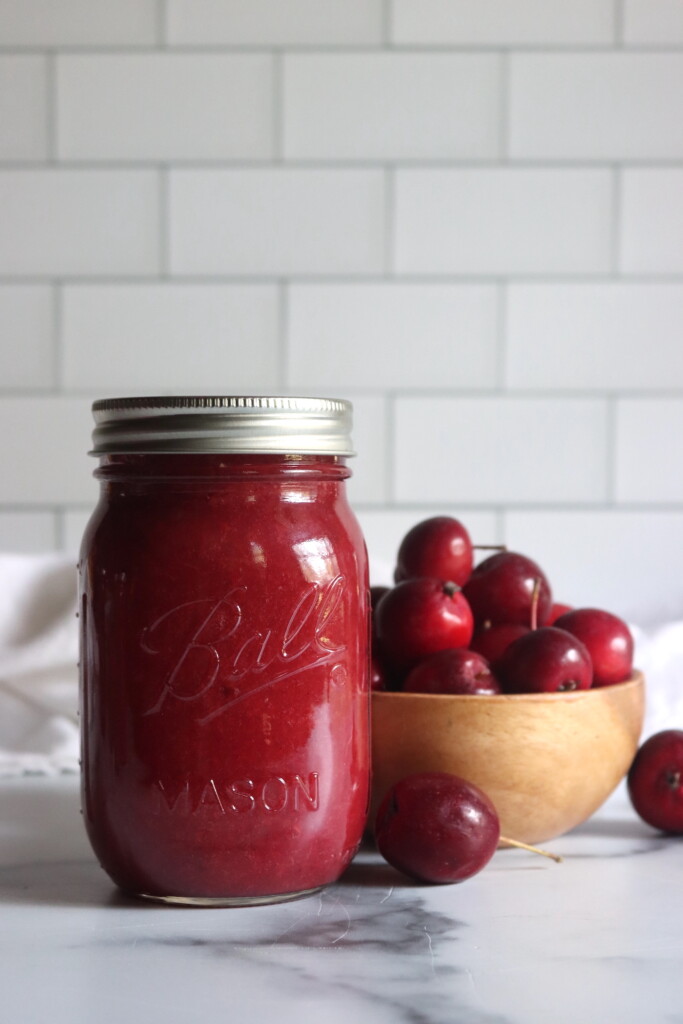 A jar of home canned crabapple sauce on a white table next to a bowl of bright red dolgo crabapples.