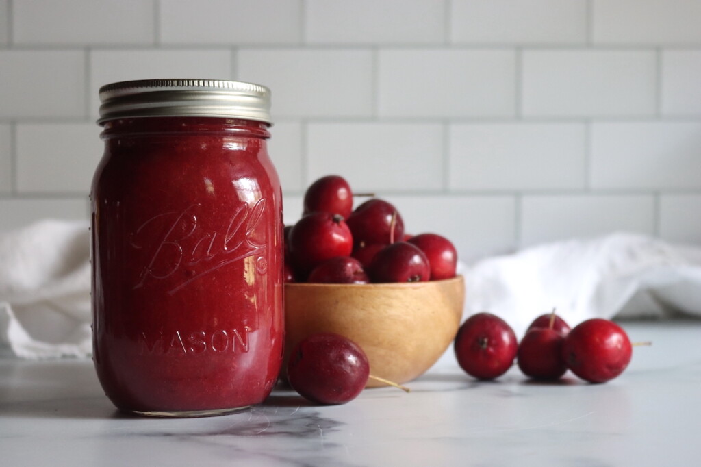 A single jar of crabapple sauce against a white background with red crabapples in a bowl nearby.