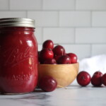 A single jar of crabapple sauce against a white background with red crabapples in a bowl nearby.