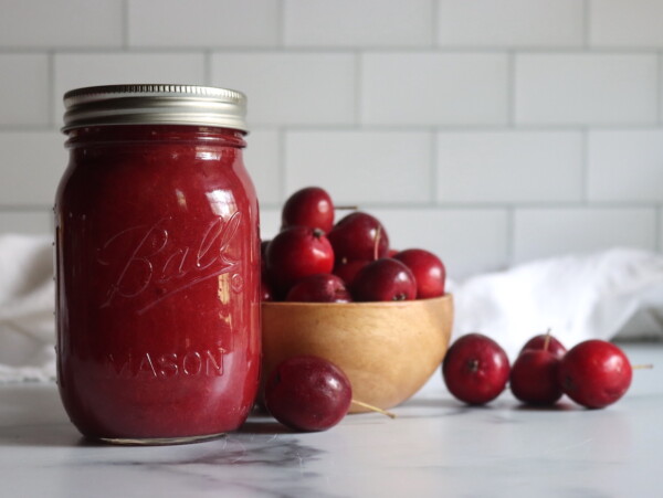 A single jar of crabapple sauce against a white background with red crabapples in a bowl nearby.