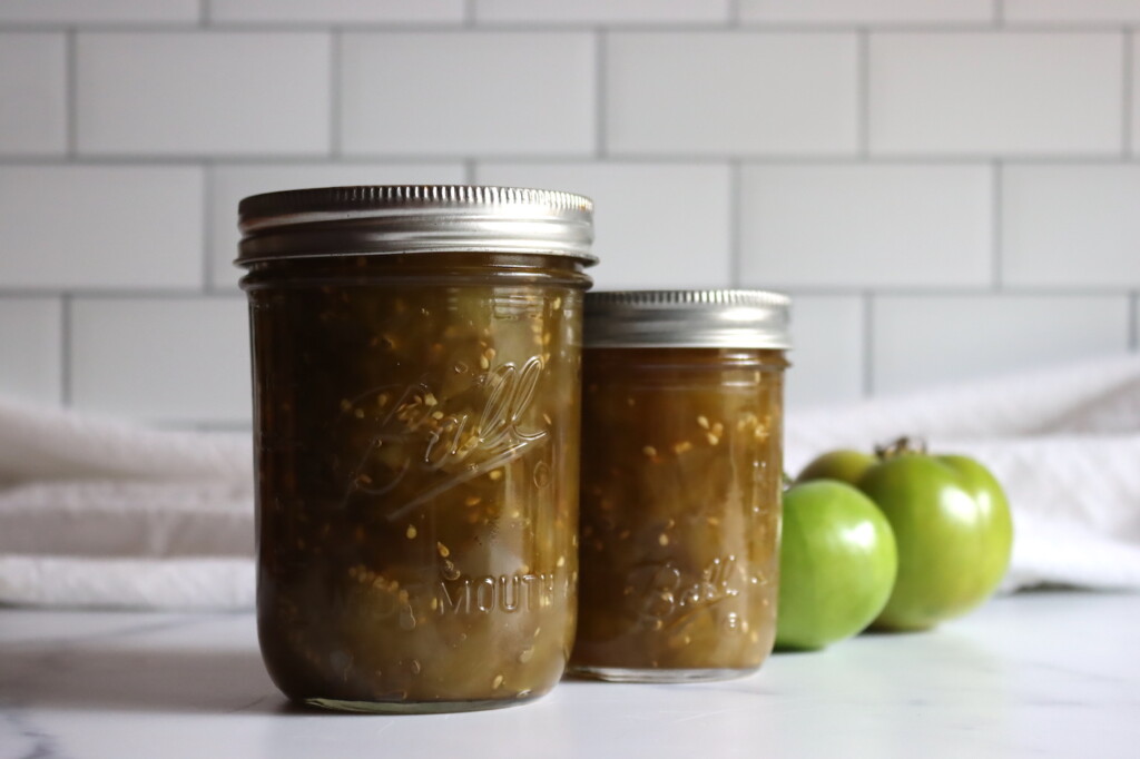 Two jars of green tomato chow chow against a white background with a few green tomatoes sitting by.