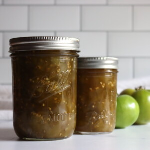 Two jars of green tomato chow chow against a white background with a few green tomatoes sitting by.
