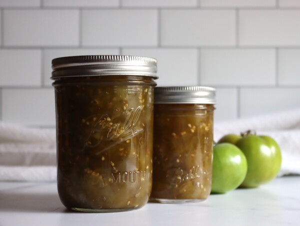 Two jars of green tomato chow chow against a white background with a few green tomatoes sitting by.