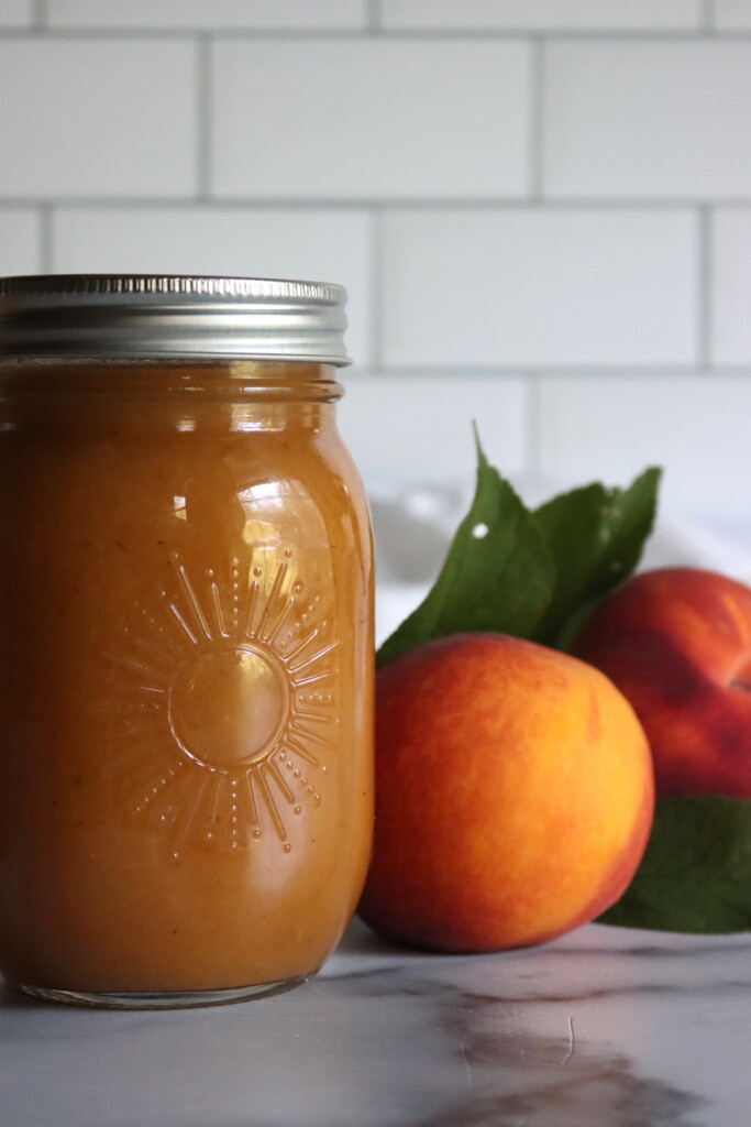 A jar of home canned peach nectar in a sunshine jar from Ball Canning, next to fresh peaches on a white countertop with a white tile background.