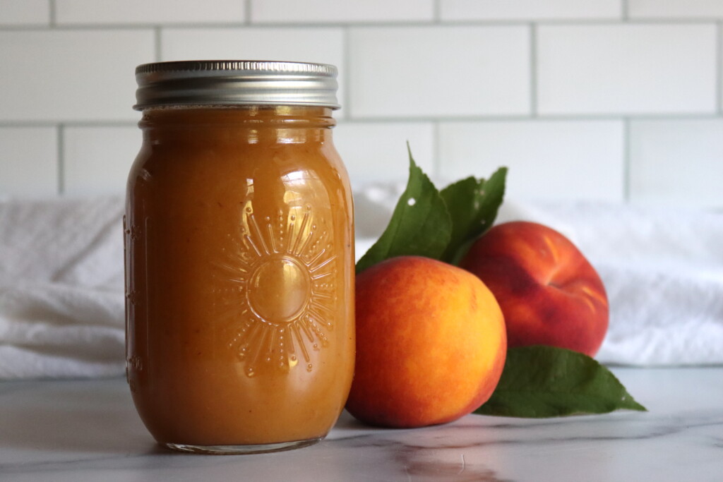 Two jars of peach nectar on a white countertop with a white tile background, with two fresh peaches next to them.