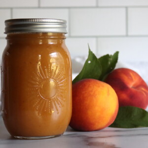 Two jars of peach nectar on a white countertop with a white tile background, with two fresh peaches next to them.