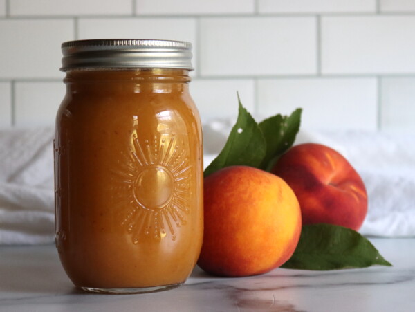 Two jars of peach nectar on a white countertop with a white tile background, with two fresh peaches next to them.