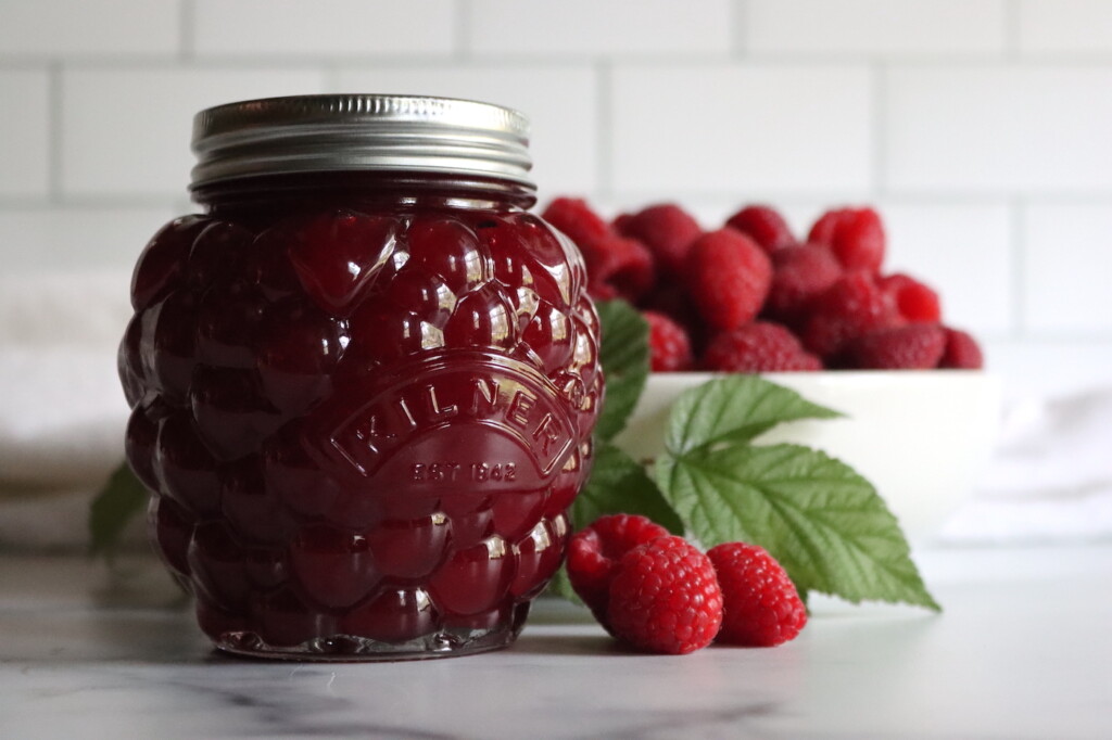 Seedless Raspberry Jam in a berry shaped jar on a white counter with a bowl of raspberries in the background.