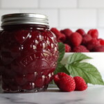 Seedless Raspberry Jam in a berry shaped jar on a white counter with a bowl of raspberries in the background.
