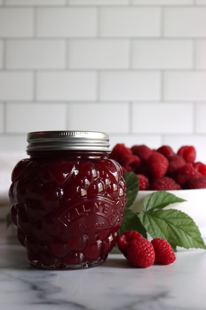 Seedless Raspberry Jam in a kilner berry jar with raspberries and raspberry leaves on a white surface.