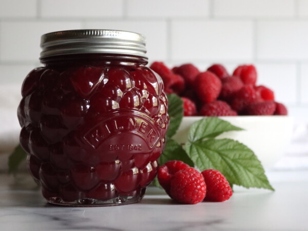 Seedless Raspberry Jam in a berry shaped jar on a white counter with a bowl of raspberries in the background.