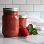 Strawberry Applesauce in a pint and half pint jar on a white table with strawberries next to it.