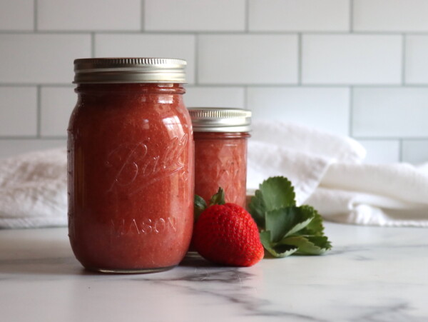 Strawberry Applesauce in a pint and half pint jar on a white table with strawberries next to it.