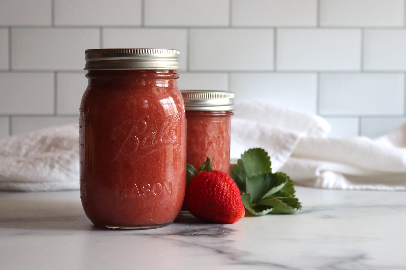 Strawberry Applesauce in a pint and half pint jar on a white table with strawberries next to it.