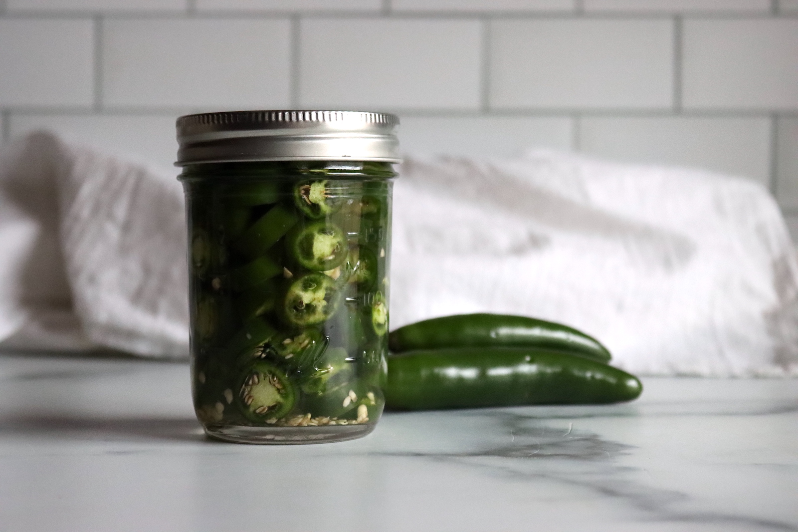 Jar of pickled serrano peppers with two fresh serranos beside it on a marble counter