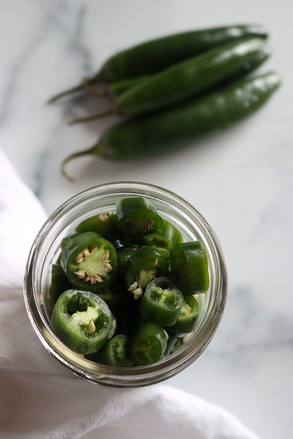 Open jar of sliced pickled serrano peppers seen from above, with fresh whole serranos beside it