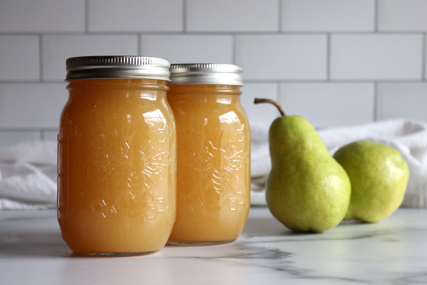 Two jars of pear honey with fresh green pears on a marble counter