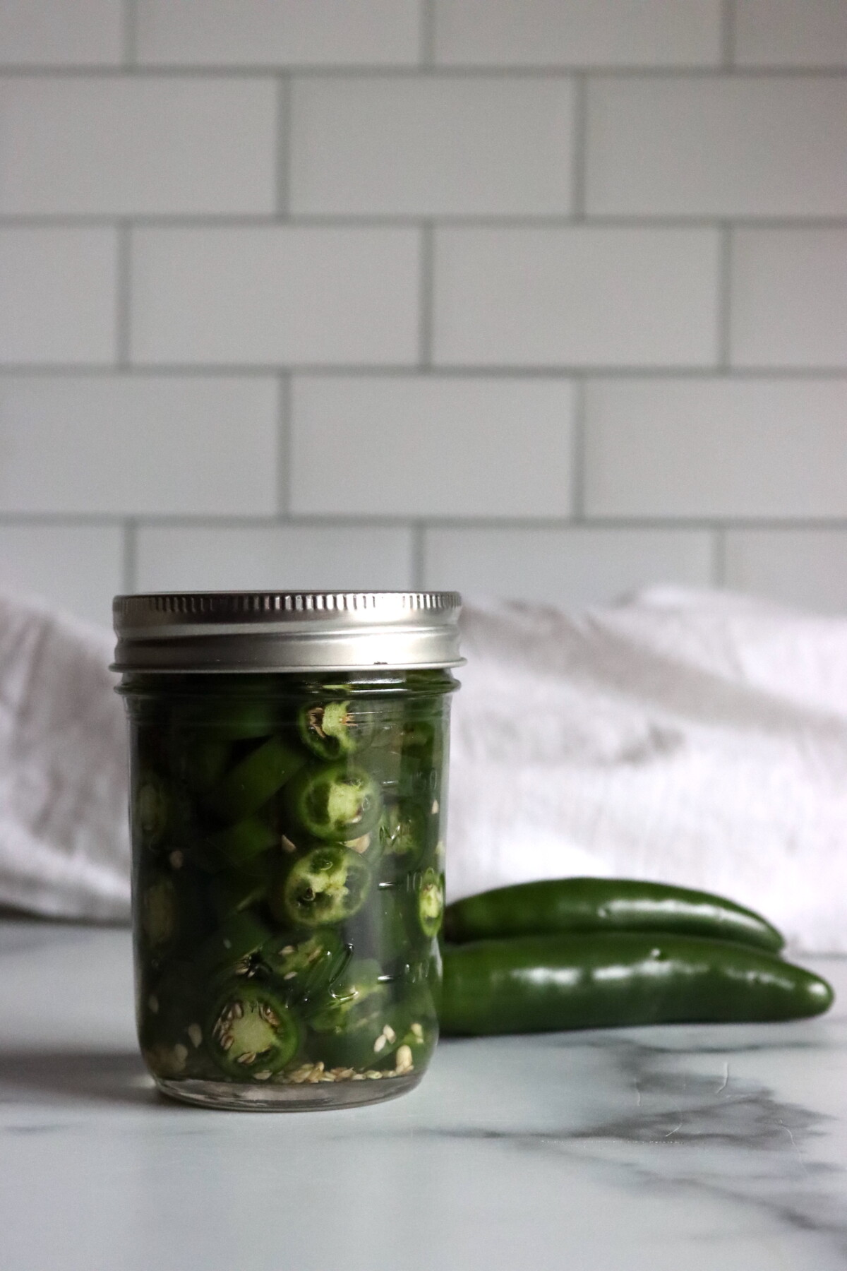 Sealed jar of pickled serrano pepper rings in brine with whole serrano peppers on the counter