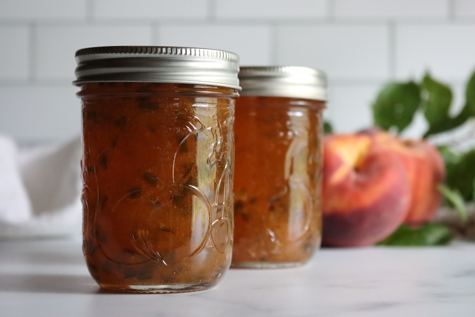 Jars of homemade peach jalapeño jam on a marble counter with ripe peaches