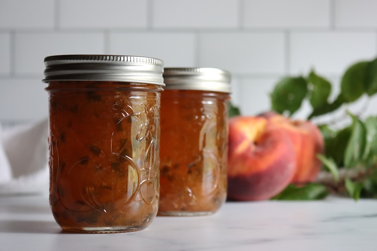 Two jars of peach jalapeño jam with fresh peaches and peach leaves