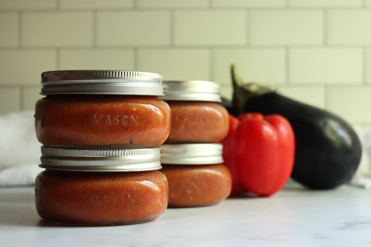 Sealed jars of canned ajvar stacked two high beside a red bell pepper and an eggplant