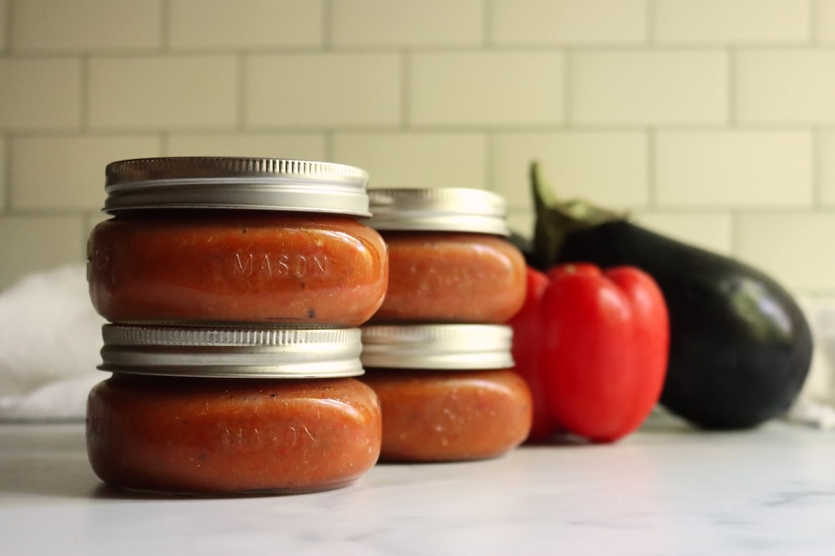 Four half-pint jars of homemade ajvar stacked in pairs, with red bell peppers and an eggplant behind them