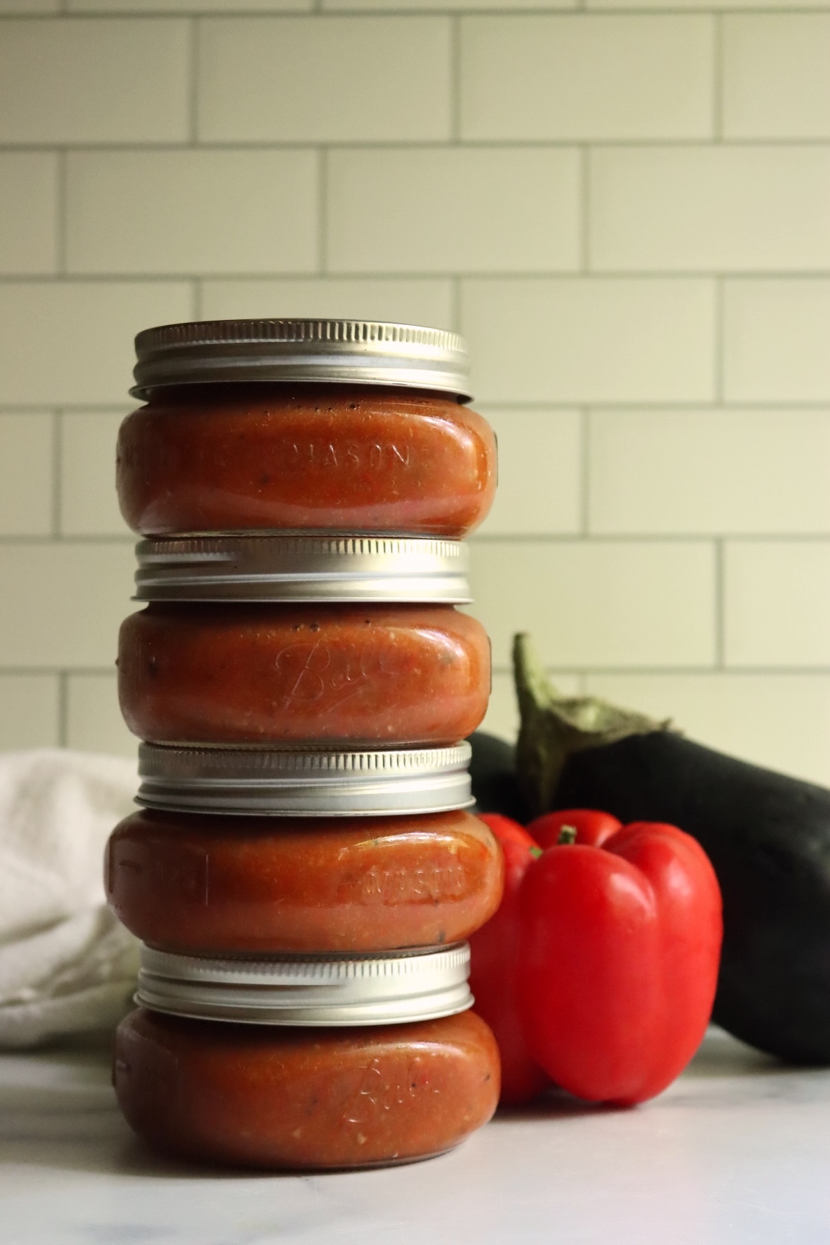 Four jars of ajvar stacked in a tower next to a red bell pepper and an eggplant