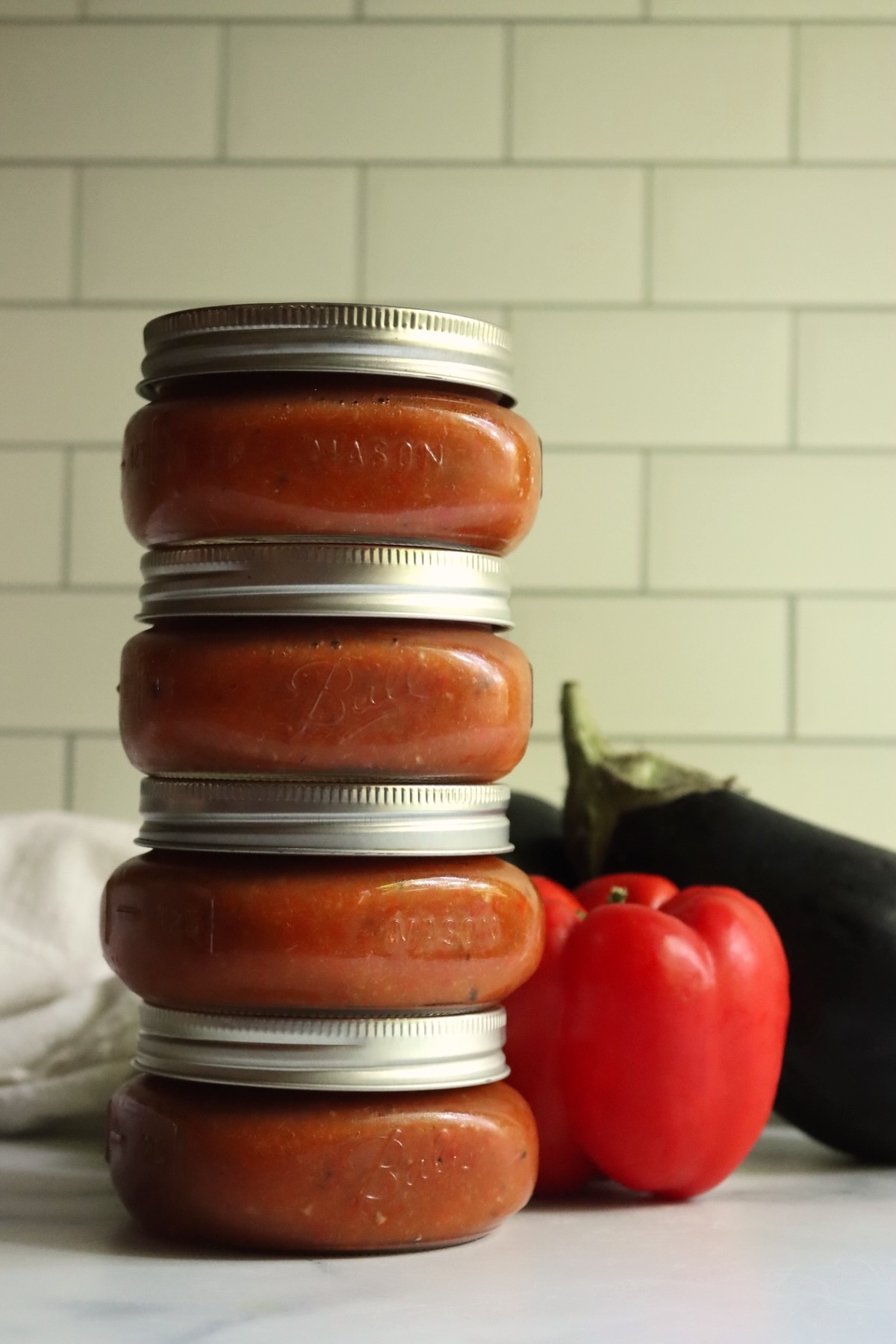 Tower of four sealed ajvar jars with a red bell pepper and an eggplant on a marble counter