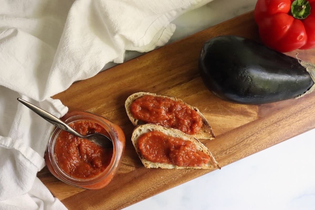 Open jar of ajvar with a spoon beside two slices of bread spread with ajvar on a wooden board