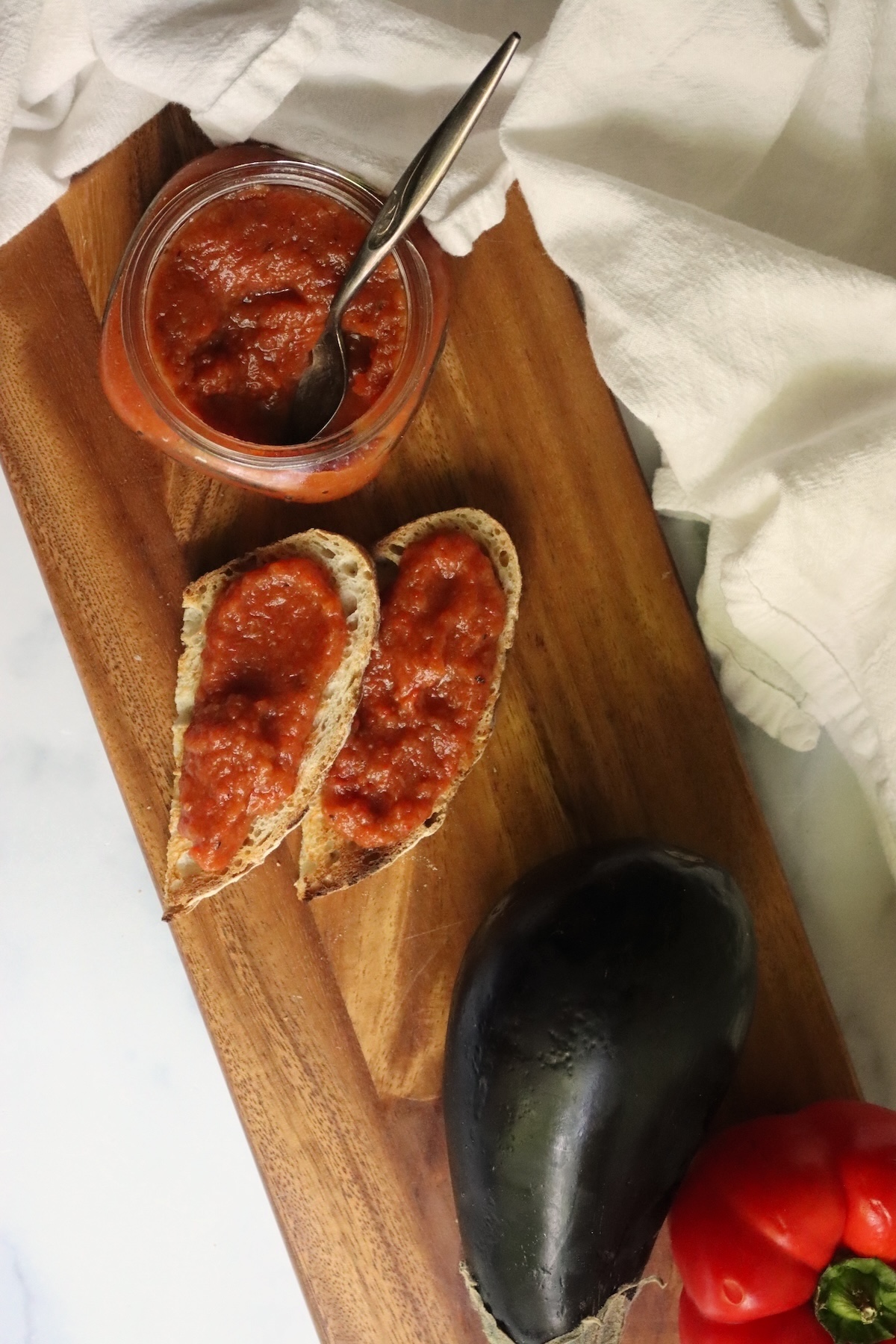 Spoon in an open jar of ajvar next to bread spread with ajvar, an eggplant, and a red pepper