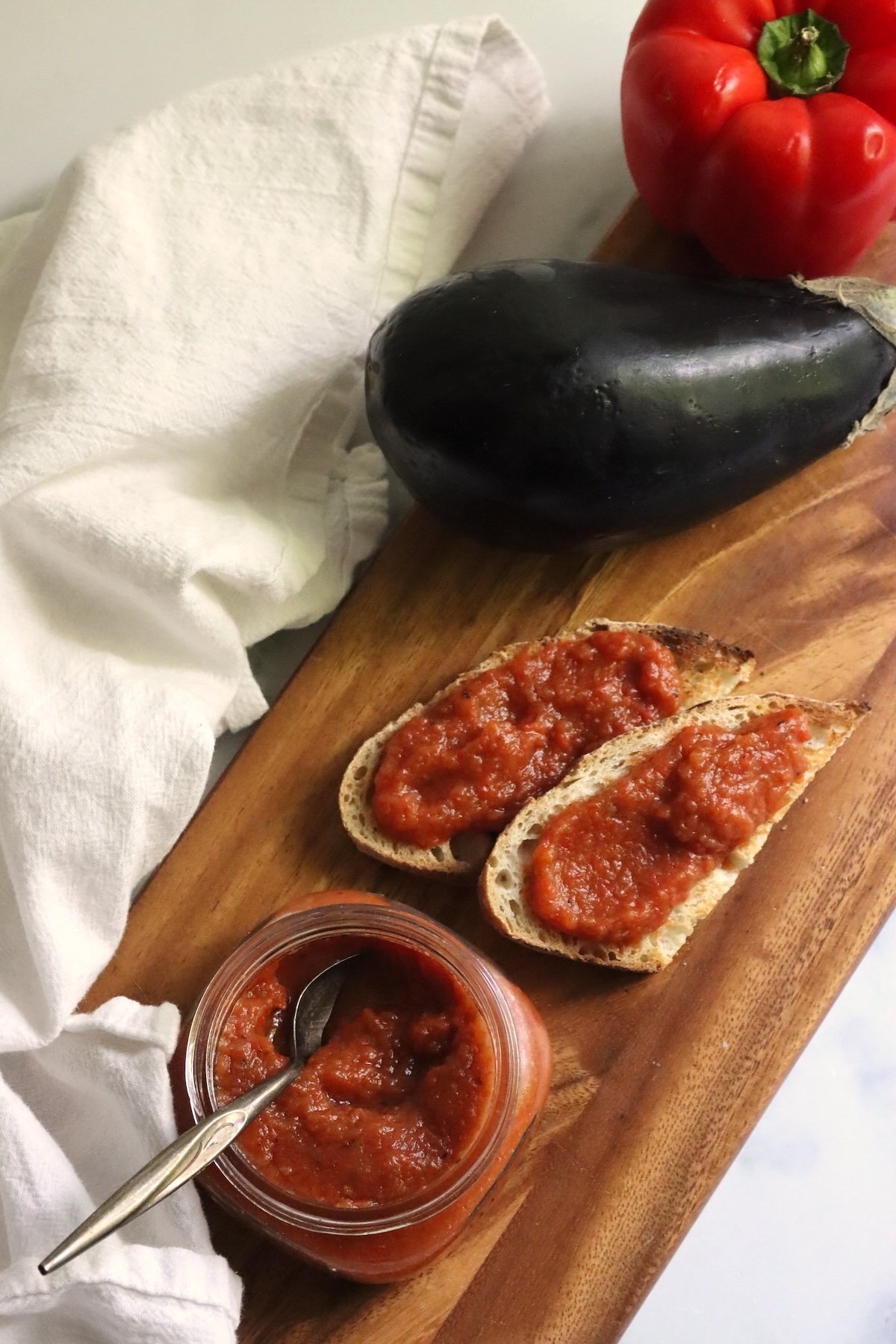 Slices of bread spread with ajvar, an open jar, an eggplant, and a red pepper on a cutting board