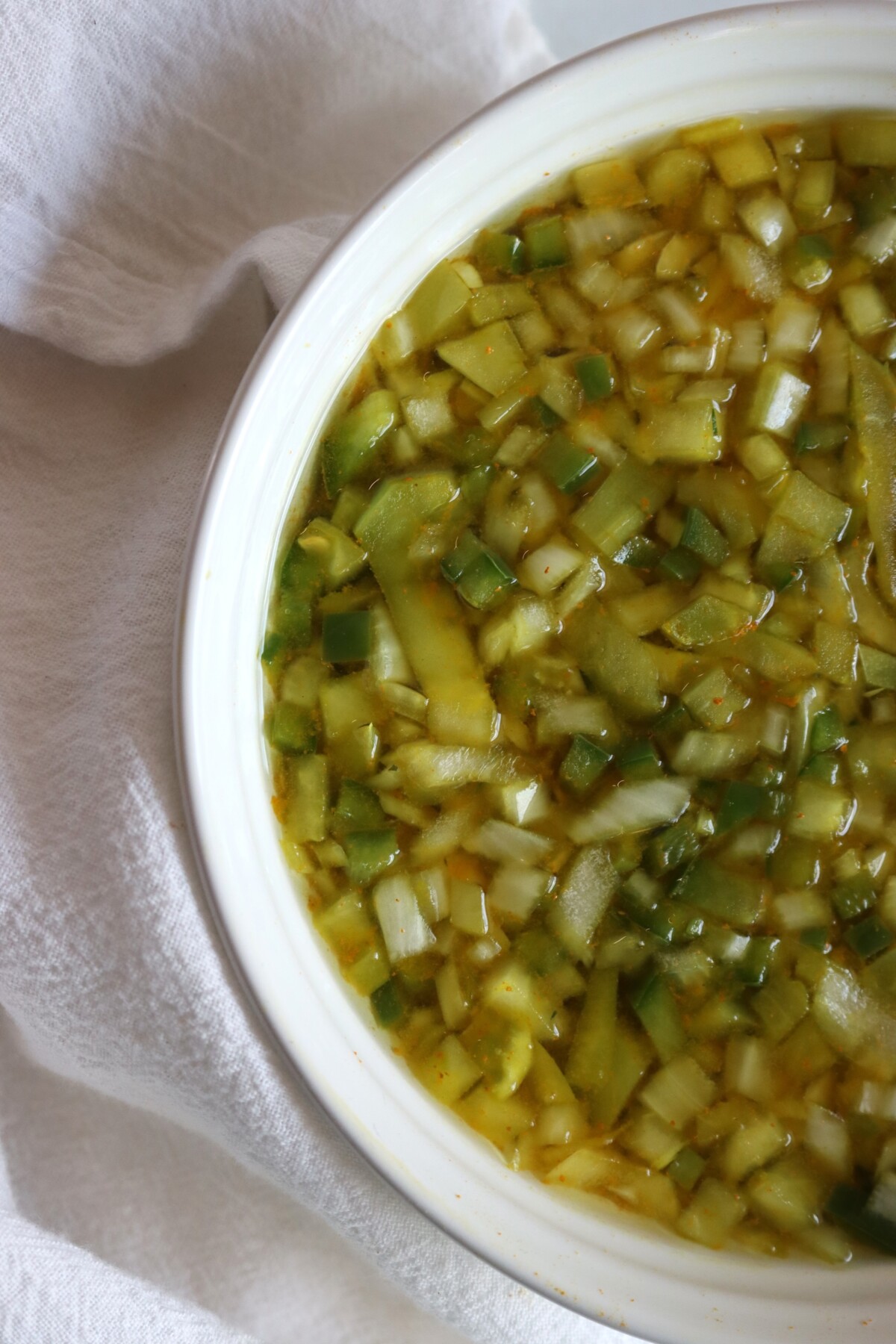 Diced green tomatoes, green peppers, and onions soaking in a salt and turmeric brine in a white bowl