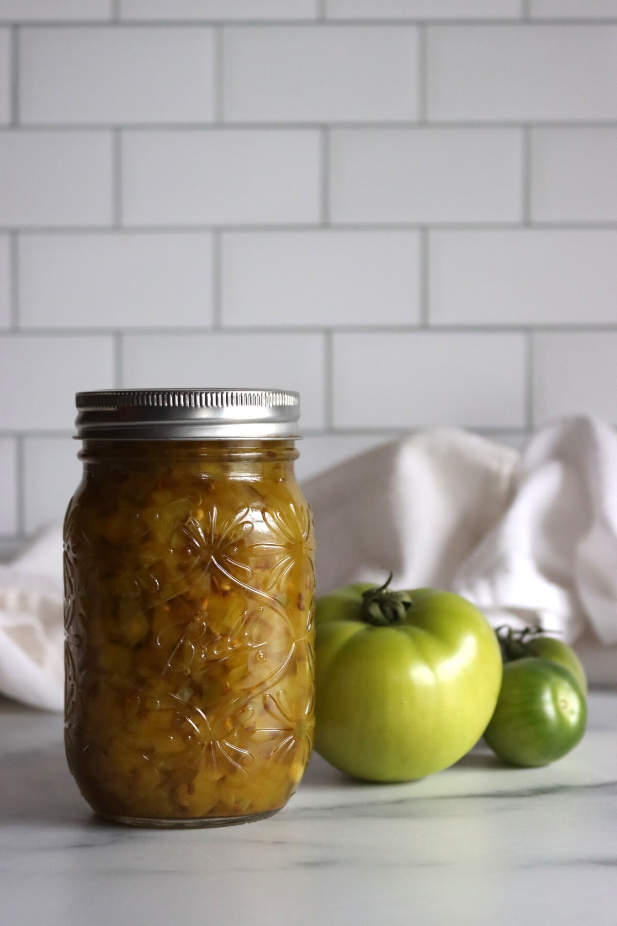Jar of dilled green tomato relish with two green tomatoes in front of a white subway tile wall