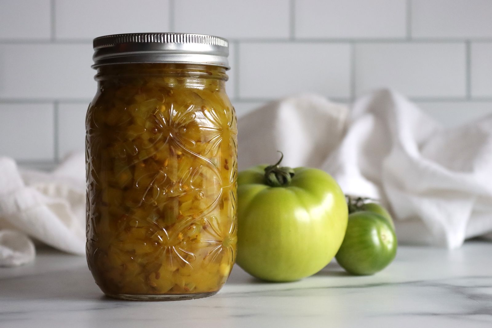 Jar of dilled green tomato relish with whole green tomatoes and a white towel on a marble counter