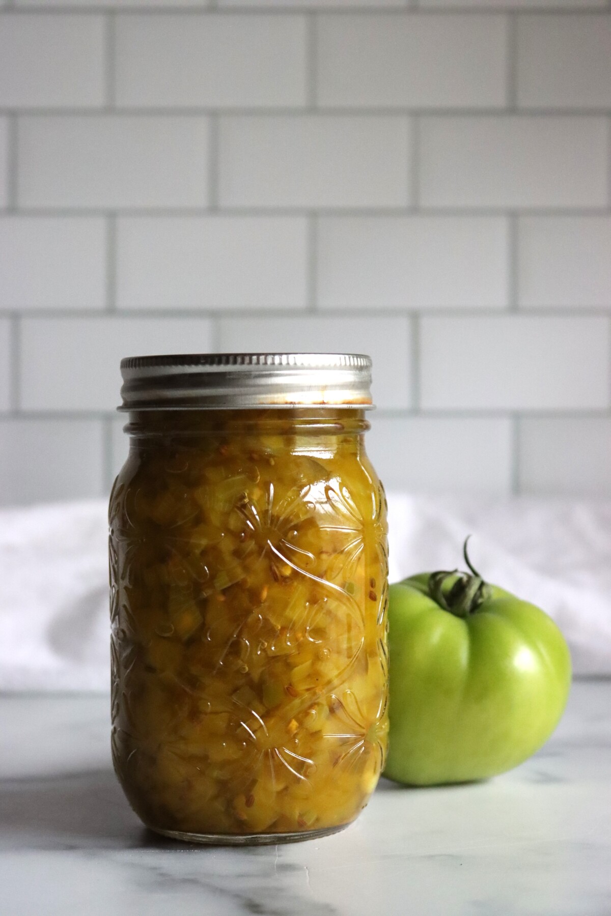 Pint jar of dilled green tomato relish beside a large green tomato in front of a white tile wall
