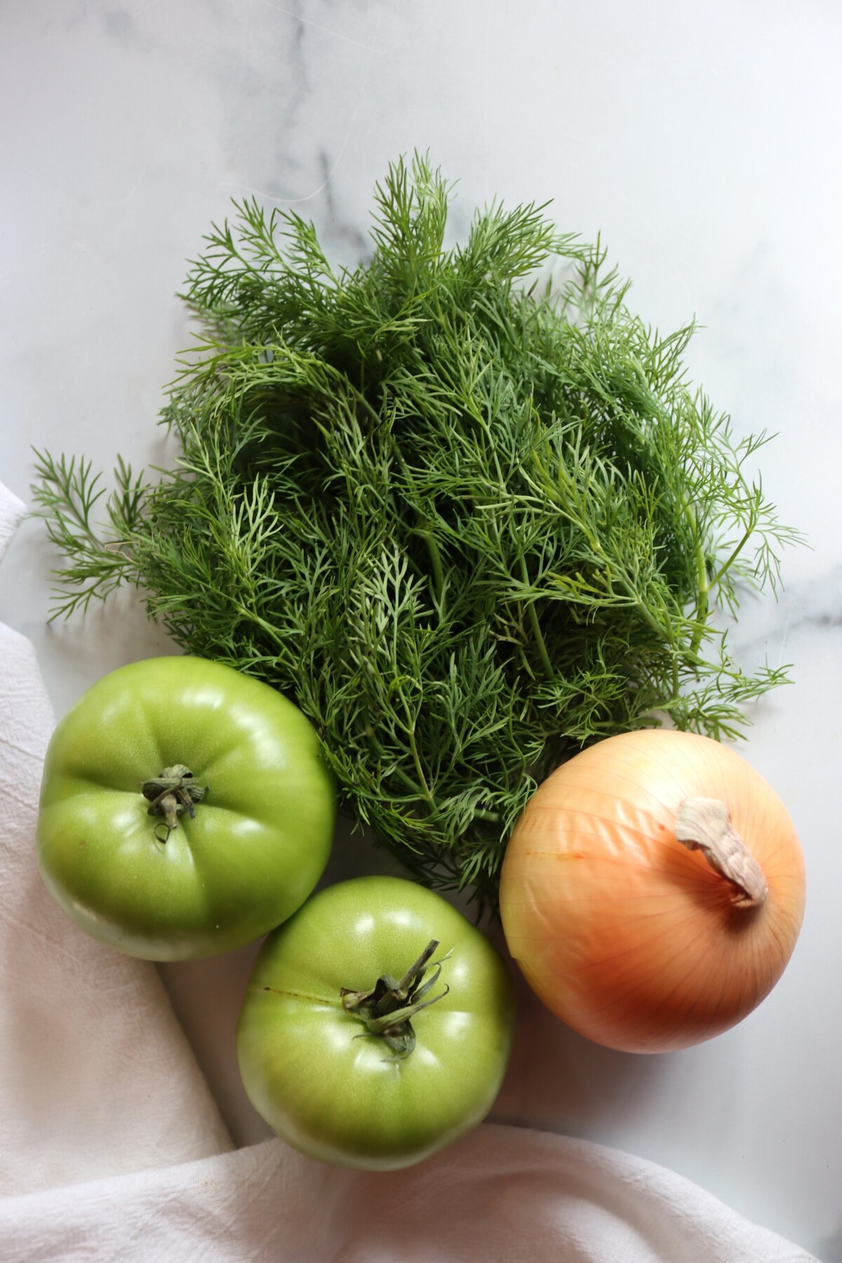 Fresh dill, green tomatoes, and a yellow onion on a white marble counter