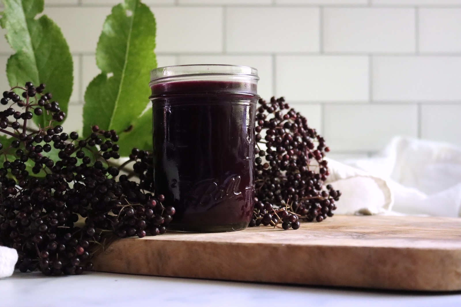 Jar of strained elderberry juice on a wooden board surrounded by fresh elderberry clusters