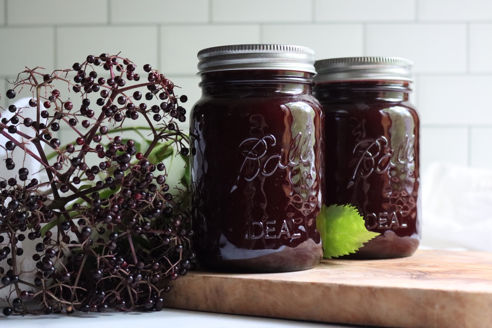 Two sealed jars of canned elderberry lemonade concentrate on a wooden board beside elderberry clusters