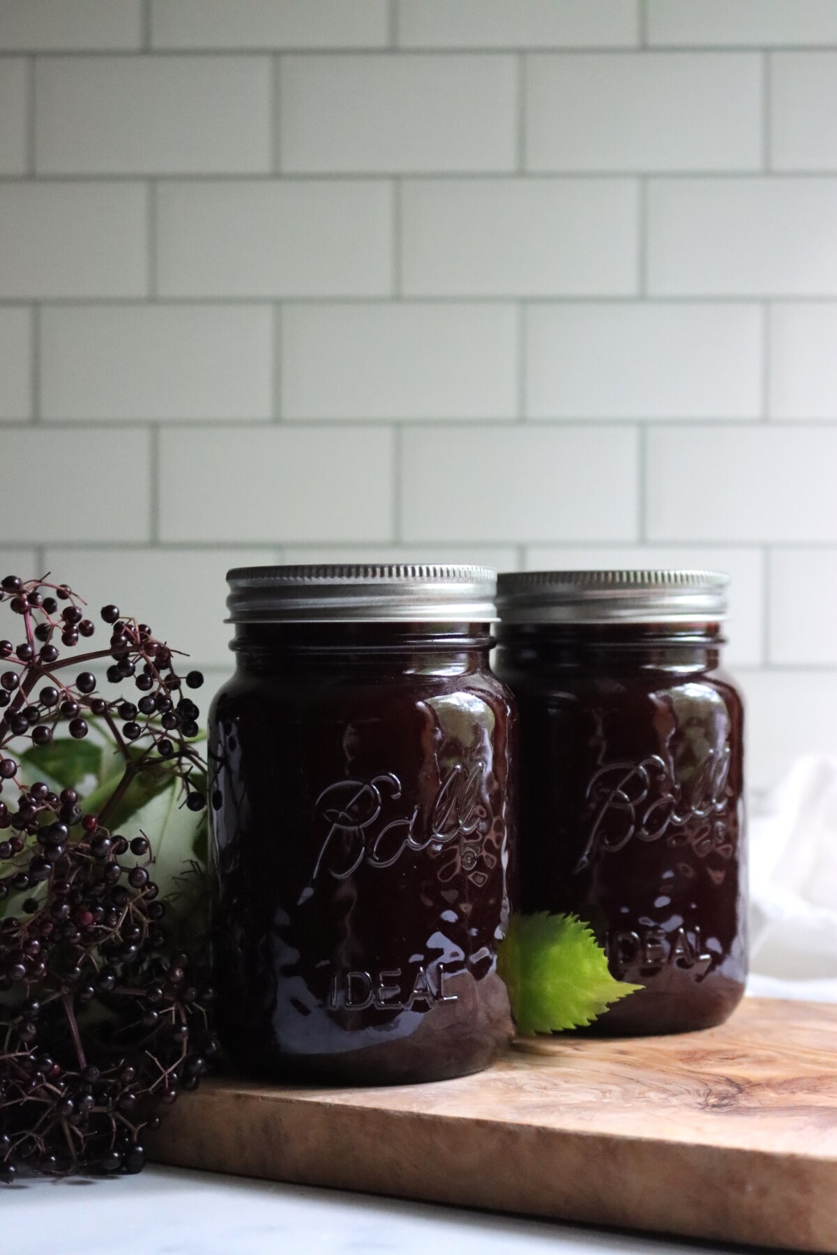Two jars of homemade elderberry lemonade concentrate on a wooden board next to fresh elderberry clusters