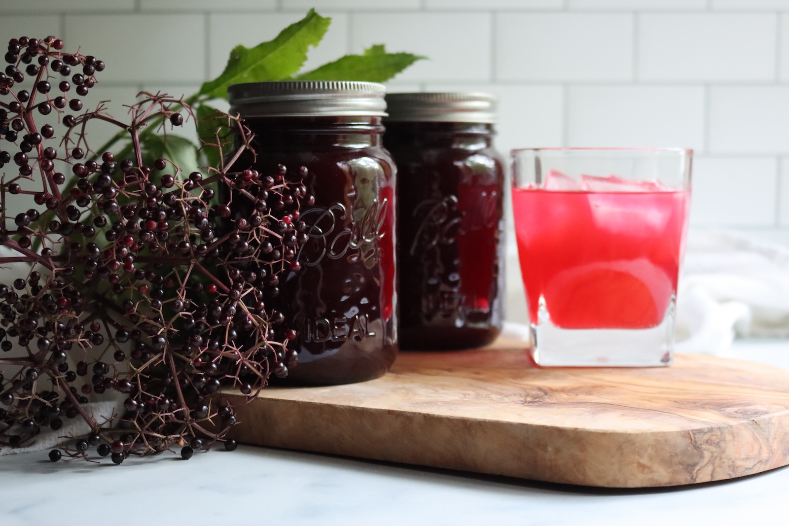 Two jars of elderberry lemonade concentrate and a glass of elderberry lemonade over ice on a wooden board