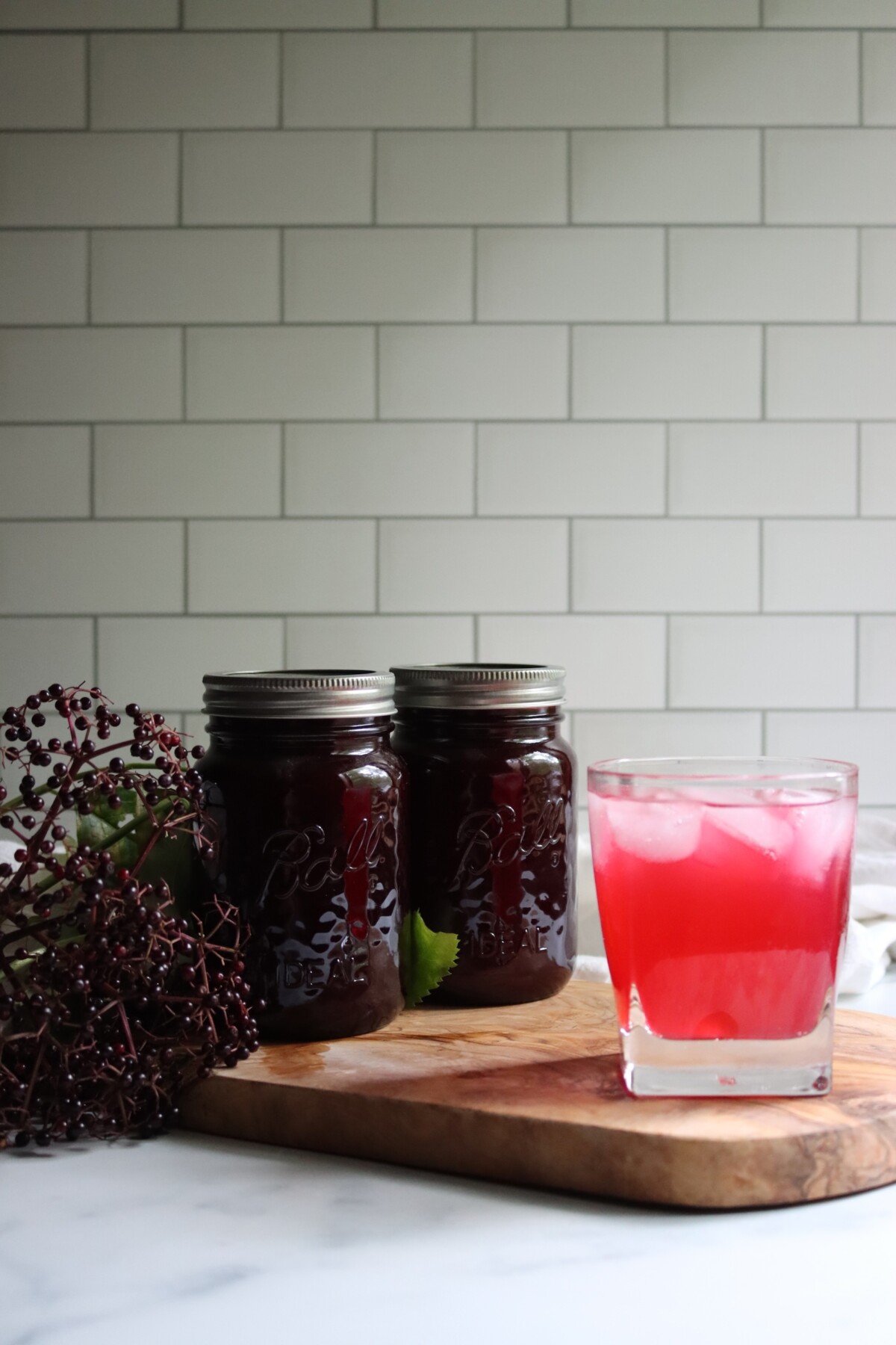 Glass of elderberry lemonade over ice beside two jars of elderberry lemonade concentrate