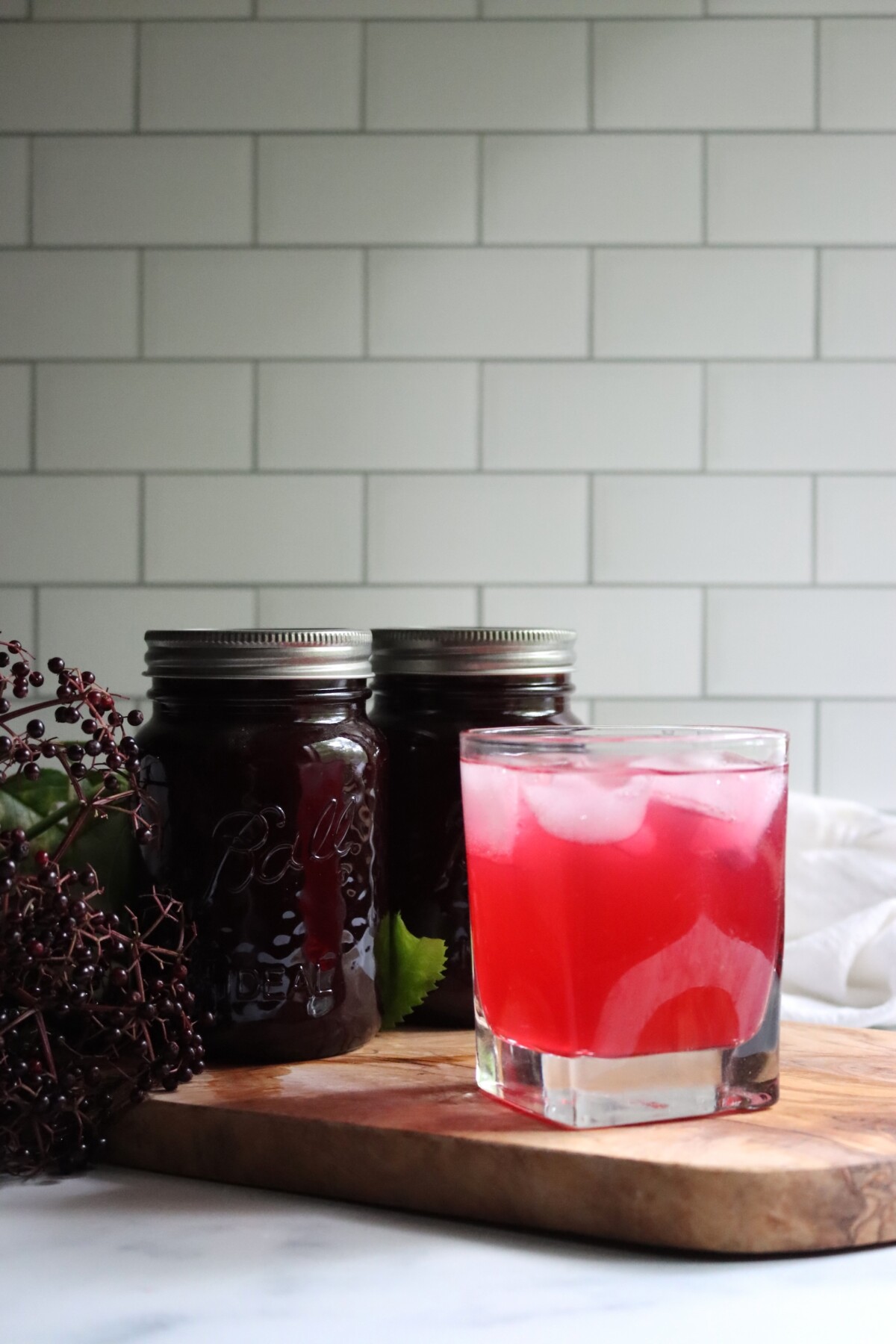 Iced glass of pink elderberry lemonade in front of two jars of concentrate on a wooden board