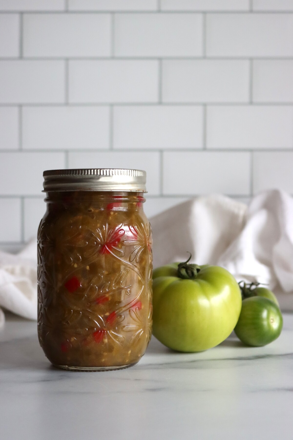 Jar of homemade green tomato hot dog relish on a counter with two green tomatoes
